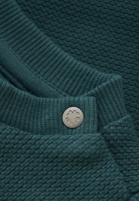 Tiefblaues Strickgewebe mit geripptem Bund und einem runden silbernen Knopf, der mit einem vierblättrigen Kleeblatt-Symbol geprägt ist, im Nahaufnahme.