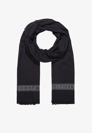 Écharpe noire Tommy Hilfiger avec une bande logo grise et bords effilochés, posée à plat avec un pli circulaire au centre supérieur.