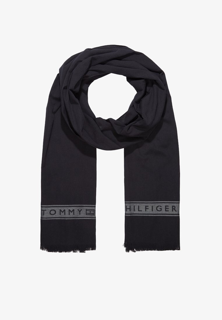 Écharpe noire Tommy Hilfiger avec une bande logo grise et bords effilochés, posée à plat avec un pli circulaire au centre supérieur.