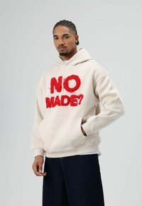 Beige hoodie met "NO MADE?" in pluizige rode letters, voorkant met kangoeroezak, capuchon met trekkoord en geribbelde boorden. Donkere spijkerbroek.