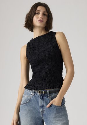 Femme aux cheveux courts bruns portant un haut sans manches noir texturé et un jean bleu clair avec une ceinture noire, main dans la poche, fond neutre.