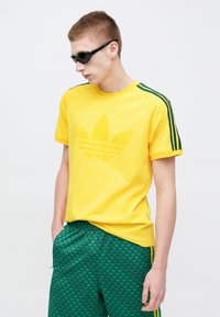 Gelbes Baumwoll-T-Shirt mit einem strukturierten grünen Blattlogo, grünen gestreiften Schulterakzenten, kombiniert mit grünen gemusterten Shorts.