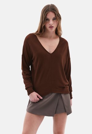 Brauner V-Ausschnitt-Pullover mit lockerer Passform, gerippten Bündchen und seitlichen Schlitzen, kombiniert mit einem knielangen grauen Faltenrock. Weiche Textur, lässiges Design.
