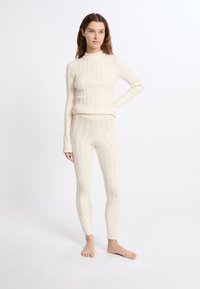 Kabelgebreide thermoset in romige kleur, bestaande uit een aansluitend langslavend top met een hoge hals en bijpassende legging met geribde boorden en een textuurpatroon.