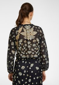 Blusa nera trasparente con motivi floreali in morbido beige e verde. Presenta maniche lunghe e una vestibilità comoda. Dettagli con vita elastica visibili.