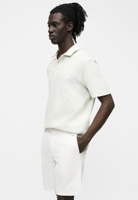 Homme avec des dreadlocks portant un polo de couleur claire et un short assorti, debout de profil avec une main dans la poche.
