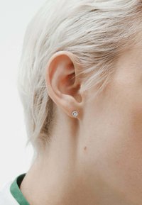 Pendientes de aro redondos con un tono plateado y una piedra preciosa clara y brillante en el centro, enmarcados por un cabello corto y de color claro.