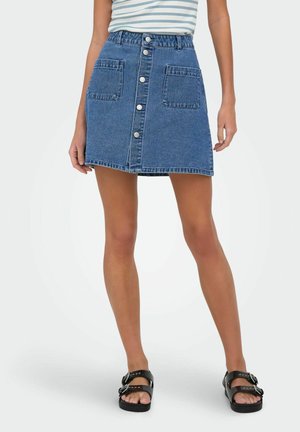Femme portant une jupe en jean bleue à boutons sur le devant avec deux poches avant, associée à des sandales noires à boucle et un haut rayé.