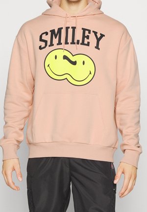 Jasnoróżowa bluza z kapturem z napisem "SMILEY" i żółtą, dwustronną grafiką uśmiechniętej buzi, noszona z czarnymi spodniami, ręce luźno wzdłuż ciała.