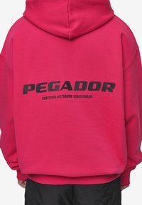 Vaaleanpunainen huppari, jossa on huppu, takana mustilla teksteillä "PEGADOR" ja "CERTIFIED AUTHENTIC STREETWEAR". Pehmeää kangasta, rento malli.