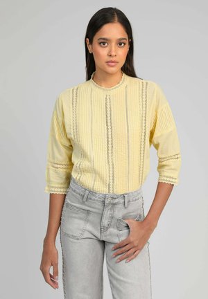 Jeune femme portant un chemisier texturé jaune clair avec des détails en dentelle et un jean denim gris clair devant un fond uni.