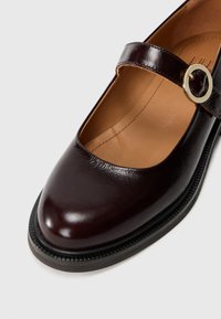 Zapato Mary Jane de cuero burdeos con puntera redonda, acabado brillante y correa con hebilla de color dorado. Suela negra texturizada con bordes elevados.
