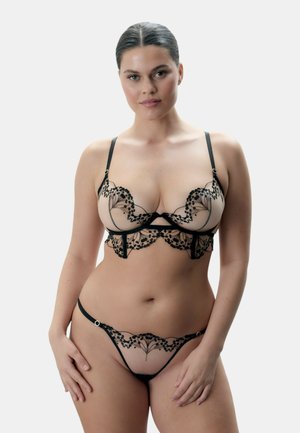 Vrouw draagt een nudekleurige en zwarte kanten lingerie set met bloemenborduursels, poserend tegen een effen lichte achtergrond.
