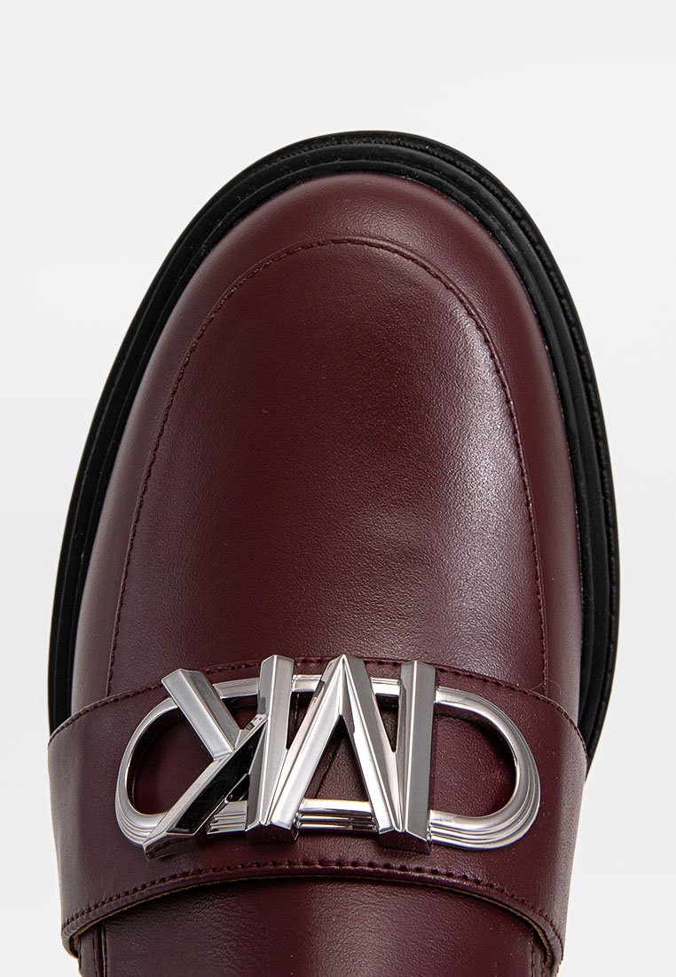 Mocassino in pelle bordeaux con un accento logato in argento lucido su una fascia, caratterizzato da una punta arrotondata e una suola in gomma nera. Texture liscia.