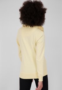 Persona con cabello rizado que lleva puesta una chaqueta de forro polar de color amarillo pálido y pantalones negros, de espaldas contra un fondo gris liso.