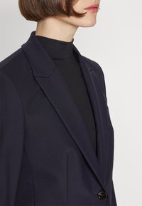 Woman wearing a dark navy blazer over a black turtleneck, shown from shoulders to chin against a plain background.