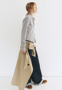 Camicia a righe, pantaloni in denim blu larghi, trench beige legato in vita, scarpe marroni e dettagli in tessuto chiaro.