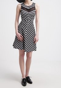 Randig klänning utan ärmar i svart och vitt med en åtsittande överdel, svängd kjol och genomskinlig panel vid halsringningen; kombinerad med svarta skor.