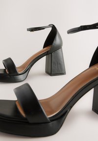 Czarne sandały na wysokim obcasie z kwadratowym noskiem i paskiem wokół kostki. Wykonane ze gładkiej skóry, z masywnym słupkowym obcasem i minimalistycznym designem.