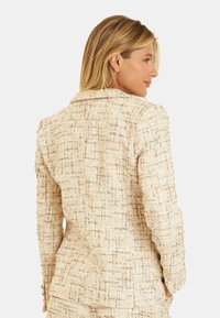 Blazer en tweed beige avec un motif texturé, design ajusté, col cranté et poignets boutonnés, mettant en avant un style structuré et sophistiqué.