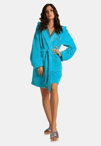 Turquoise hooded robe made of plush material, featuring a tie closure and front pockets, worn over patterned slide sandals.