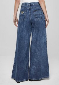 Mėlyni platėjančių kojų džinsai iš medvilninio audinio, turintys išblukusį atspalvį, aukštą liemenį ir du galinius kišenes su logotipo etikete. Paprastas paviršius be matomų dekoracijų.