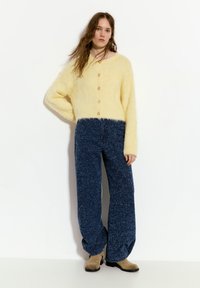 Gelber flauschiger Cardigan mit einem runden Ausschnitt und braunen Knöpfen, kombiniert mit lockeren blauen strukturierten Hosen. Das Modell steht vor einem weißen Hintergrund.