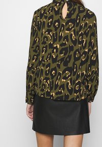 Blusa verde con stampa leopardata e dettagli neri, dotata di uno scollo a chiave sul retro, maniche lunghe e una vestibilità rilassata, abbinata a una gonna in pelle nera.