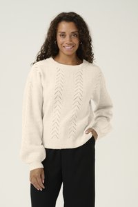 Krämfärgad stickad tröja med strukturerade sömmar och puffärmar, bärs över svarta byxor, med rund halsringning och öppna mönster.