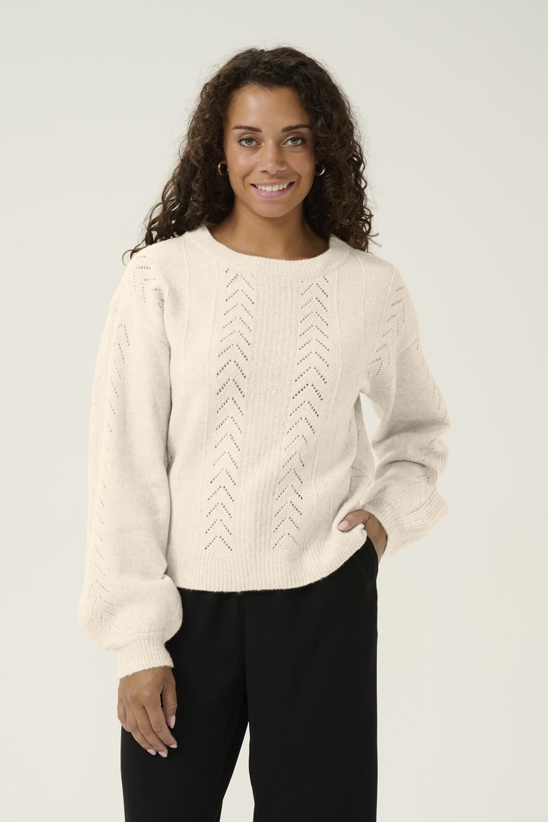 Krämfärgad stickad tröja med strukturerade sömmar och puffärmar, bärs över svarta byxor, med rund halsringning och öppna mönster.