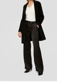 Manteau noir, pull à col en V crème et pantalon en satin noir taille haute avec détail noué. Le mannequin porte des bottines noires à talons.