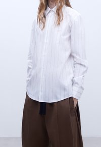 Camicia bianca con bottoni, a righe verticali sottili, maniche lunghe e colletto a punta. Vestibilità comoda, abbinata a pantaloni larghi marroni.