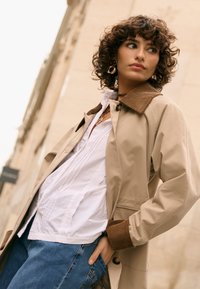 Kobieta o kręconych włosach, ubrana w beżowy płaszcz z brązowym kołnierzem, białą bluzkę, niebieskie jeansy oraz czerwono-złote kolczyki, stoi na zewnątrz przy budynku.