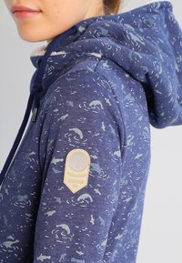 Bluza z kapturem w granatowym kolorze z wzorem pływających zwierząt. Zawiera teksturowaną beżową naszywkę z logo na rękawie.