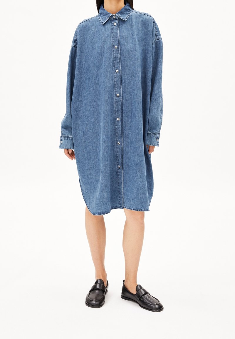 Femme portant une robe chemise en denim bleue oversize, associée à des mocassins en cuir noir, debout devant un fond blanc.