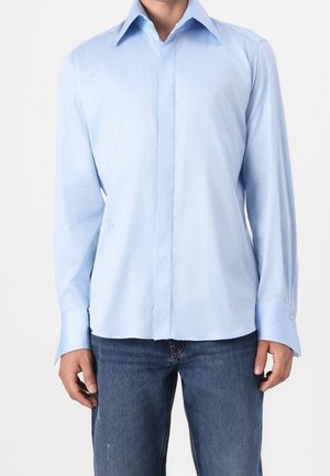 Mężczyzna w jasnoniebieskiej koszuli z długim rękawem zapinanej na guziki oraz niebieskich dżinsach, stojący na jednolitym białym tle.