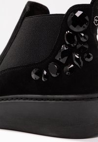 Černé klouzací boty s textilním svrškem a texturovaným povrchem, zdobené černými dekoračními kameny na boku. Plochá gumová podrážka s detailem šití.