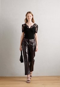 Haut noir avec des manches courtes en dentelle, associé à un pantalon en cuir brun foncé et des sandales à lanières. Le mannequin tient un petit sac noir.
