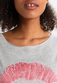 Nainen, jolla on kihara hius, pukeutuneena harmaaseen paitaan, jossa on iso tekstuuroitu vaaleanpunainen huulipainatus etupuolella, kuvattuna huulista ylös rintakehään.