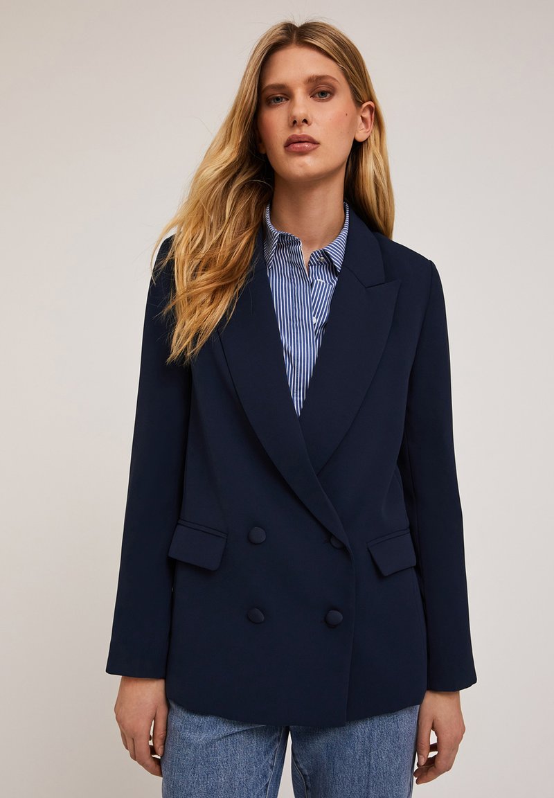 Granatowa marynarka dwurzędowa z klapami i dwoma przednimi kieszeniami, zestawiona z jasnoniebieską koszulą w prążki. Gładka tekstura materiału.