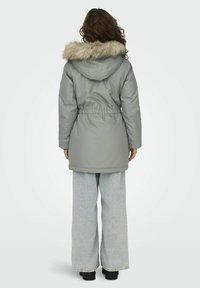 Parka grise en ouatiné avec une capuche en fausse fourrure, taille avec cordon de serrage, et longueur mi-cuisse. Associée à un jean ample bleu clair et des bottines noires.