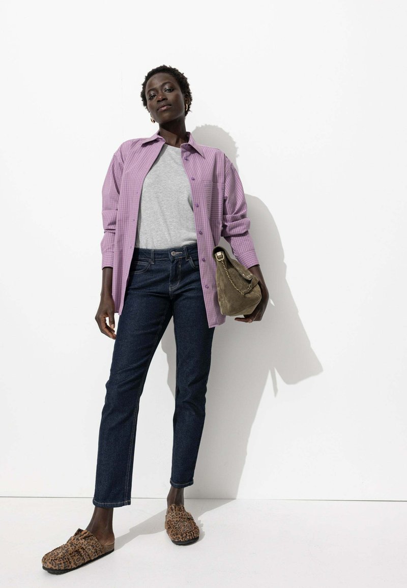 Chemise à carreaux violette avec un t-shirt gris en dessous, associée à un jean slim noir et des chaussures à enfiler marron avec imprimé léopard, tenant un sac à main vert.