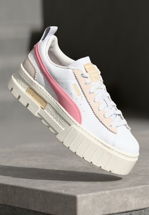 White and beige Puma sneaker with pink stripe, thick textured sole, and laces, displayed mid-air above concrete surface.