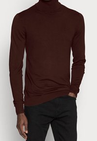 Maglione a collo alto marrone, con colletto e polsini a coste, design aderente, texture morbida, indossato con pantaloni neri.