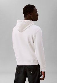 Mann sett fra siden iført en ensfarget hvit hettegenser med hetten nede og mørke jeans mot en grå bakgrunn.