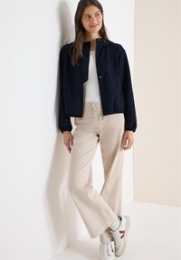 Veste bleu marine zippée avec col arrondi, poignets élastiques et poches, associée à un pantalon ample beige clair et des baskets multicolores.