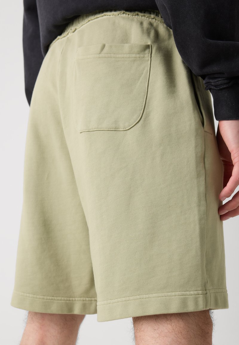 Hellgrüne Baumwollshorts mit einem elastischen Bund, seitlichen Taschen und einer einzigen Gesäßtasche. Glatte Textur, knielanger Schnitt.