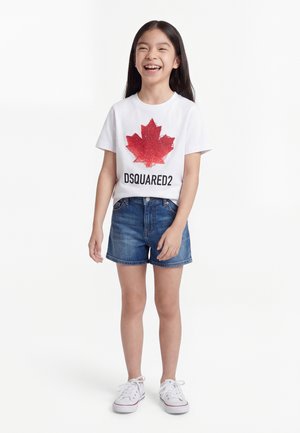 Fille souriante debout, portant un t-shirt blanc avec une feuille d'érable rouge et le texte "DSQUARED2", un short en denim et des baskets blanches.