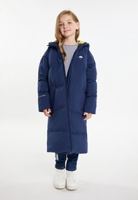 Cappotto imbottito blu navy con cappuccio, dotato di fodera gialla, chiusura con zip e logo. Indossato sopra una maglietta grigia e jeans di denim. Sneakers bianche.