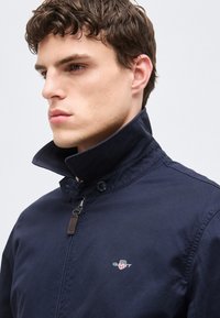 Noor mees lühikeste lokkis juustega, kes kannab tumesinist lukuga jope tõstetud kraega ja väikese GANTi logoga rinnal.